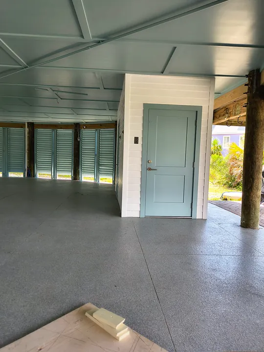Ground level under a stilt beach house with painted ceiling, white lap siding enclosure and a blue-gray door