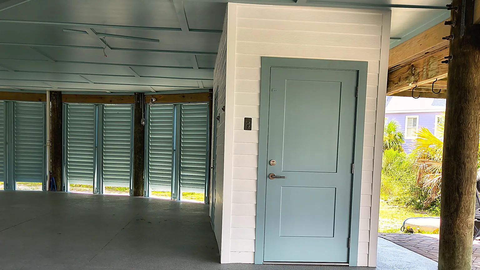 Ground level under a stilt beach house with painted ceiling, white lap siding enclosure and a blue-gray door